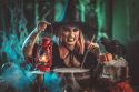 Witch with with awfully face and lighted lantern in creepy surroundings cooks poison soup in boiling cauldron.